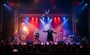 Circus performers on stage: aerialist, fire breather, contortionist, and hosts in costume, under colorful stage lights.