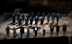 A group of performers in historical costumes stand holding hands on a stage.