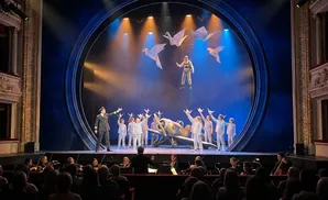 A performer in mid-air, suspended by strings from origami birds, above a stage with children, a prop airplane, and an orchestra.