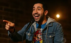 A man with a beard points while laughing into a microphone, wearing a denim jacket over a Mario t-shirt.