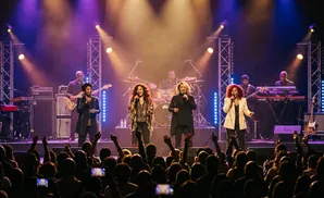 A female vocal group performs on stage with a band under purple lights as an audience cheers.