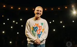 Man with short blonde hair smiles in a white sweater with colorful hearts, standing on a dark stage with lights.