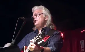 Musician with long gray hair and glasses playing guitar on stage.