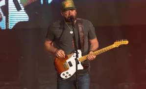 Lee Brice performs on stage, wearing a green hat and playing a guitar.