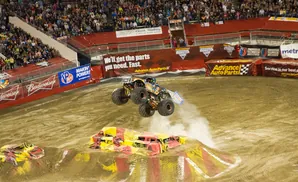 A monster truck leaps over crushed cars in a dirt arena with a large crowd watching.