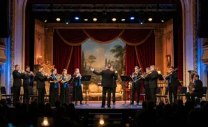 An orchestra performs on a stage with red velvet curtains and ornate gold trim.