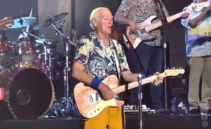 Man in a Hawaiian shirt plays a light-colored electric guitar on stage, with a drum set and other musicians in the background.