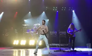 A Freddie Mercury impersonator, shirtless in white pants, performs on stage with a band.