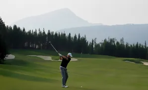 Golfer mid-swing on a lush green course with a line of pine trees and misty mountains behind.