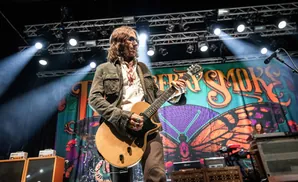 A guitarist on stage, illuminated by spotlights, plays a wooden electric guitar in front of a colorful butterfly backdrop.