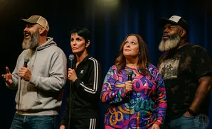 Four comedians stand on stage holding microphones, looking upwards.