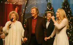 A group of singers perform on stage with Christmas trees in the background.