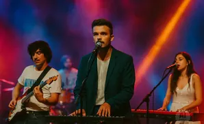 A band performs on stage with a male singer in the foreground, a guitarist to his left, and a female keyboardist to his right.