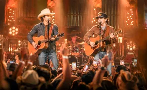 Two male country singers with guitars perform on stage, with a cheering crowd holding up phones in the foreground.