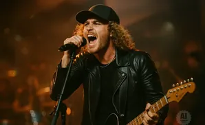 A man with curly hair wears a black leather jacket and a baseball cap while singing into a microphone and playing a guitar.
