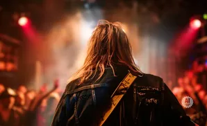 Musician with long, brown hair from behind, wearing a denim jacket on a dimly lit stage with a cheering crowd.