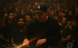 A DJ in a black cap and shirt plays music for a crowd in a dimly lit club.