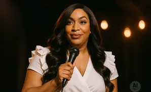 A woman with long dark hair and a white ruffled top holds a microphone and smiles at the camera.