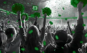 A crowd cheers at an event, raising green pom-poms and throwing green confetti.