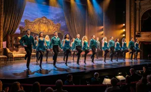 Dancers in teal costumes perform on stage with a theatrical backdrop and audience in the foreground.