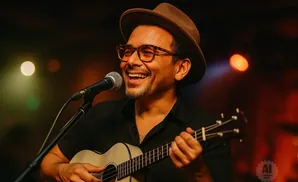 A smiling man in a hat and glasses sings into a microphone while playing a ukulele.