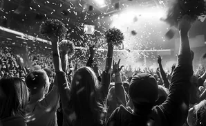 Black and white image of a cheering crowd at a concert, with confetti falling and hands raised holding pom-poms.
