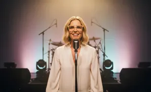 A smiling woman with blonde hair and glasses stands behind a vintage microphone on a stage, with a drum set in the background.