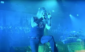 Man with long hair and beard, wearing dark clothing and holding a microphone, performs on stage with blue lighting and a Viking-themed backdrop.
