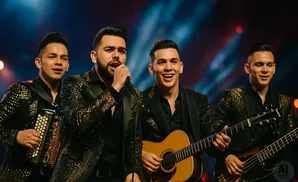 Four male musicians in sparkly jackets perform on stage, with one singing into a microphone, one playing an accordion, one playing an acoustic guitar, a…