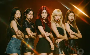 Five K-pop singers on stage, wearing matching black outfits and denim shorts, with one singer in red hair.