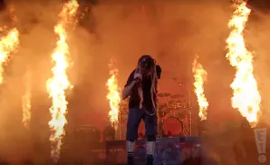 A performer with dreadlocks sings on a stage surrounded by tall flames.