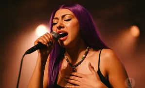 A woman with purple hair sings into a microphone while holding her chest.