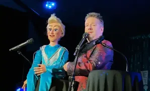 A ventriloquist performs with a blonde puppet wearing a blue dress, both in front of microphones on a stage.
