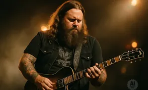 A long-haired, bearded man with tattoos plays an electric guitar on stage with warm lighting.