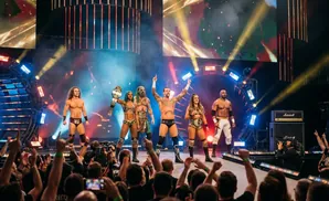 Wrestlers in ring gear stand on stage with belts, cheered on by a crowd.