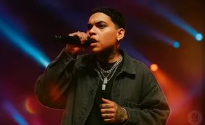 A person with tattoos and jewelry sings into a microphone on stage, illuminated by stage lights.