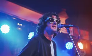 A male singer with dark, curly hair and white sunglasses sings into a microphone on a stage lit by blue and pink lights.