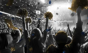 A crowd cheers and throws confetti during a celebration, with some holding gold pom-poms.