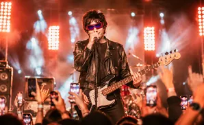 A rock singer in a leather jacket and sunglasses performs on stage, playing a bass guitar in front of a crowd with phones raised.