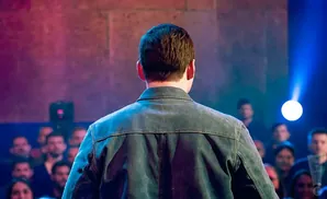 Man in denim jacket facing away from camera, on stage with audience in background.