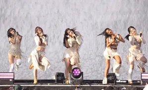 Five women in light-colored outfits dance on a stage in front of a large, bright screen.