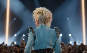 Back view of a performer with blonde hair, wearing a light blue blazer with sparkly shoulder pads, on stage in front of a crowd.