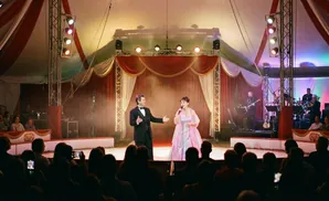 Performers in a circus tent, a man in a tuxedo and a woman in a gown, sing for an audience.