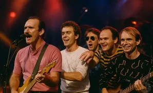 A band performs on stage with five men singing and playing instruments.