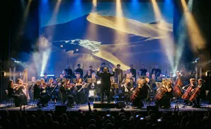 An orchestra performs live on stage with a large screen displaying a desert landscape with a spaceship.