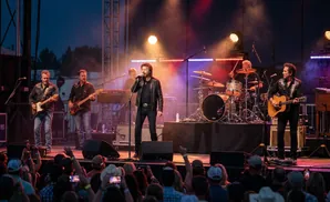 A country band performs on a brightly lit stage at night for a large, enthusiastic crowd wearing cowboy hats.