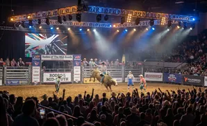 A cowboy rides a bucking bull in a rodeo arena, with a crowd watching and lights shining down.