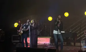 A band performs on stage, with singers at microphones and a guitarist.