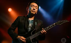 A guitarist in a suit plays a black electric guitar under stage lights.