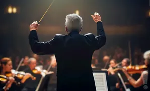 A conductor with gray hair leads an orchestra, holding a baton and gesturing with both hands.